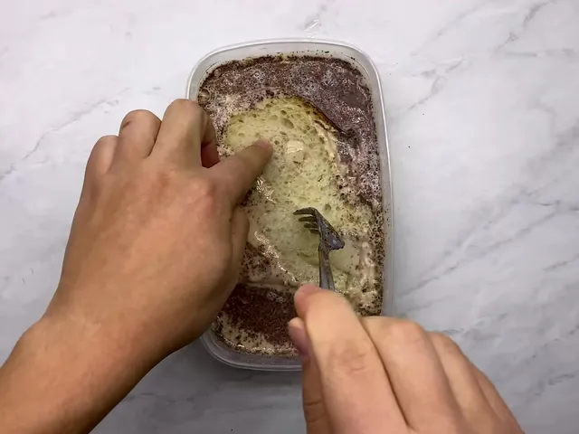 Hands pressing a thick slice of sourdough into the egg, milk and cinnamon mixture in a plastic container, with a fork