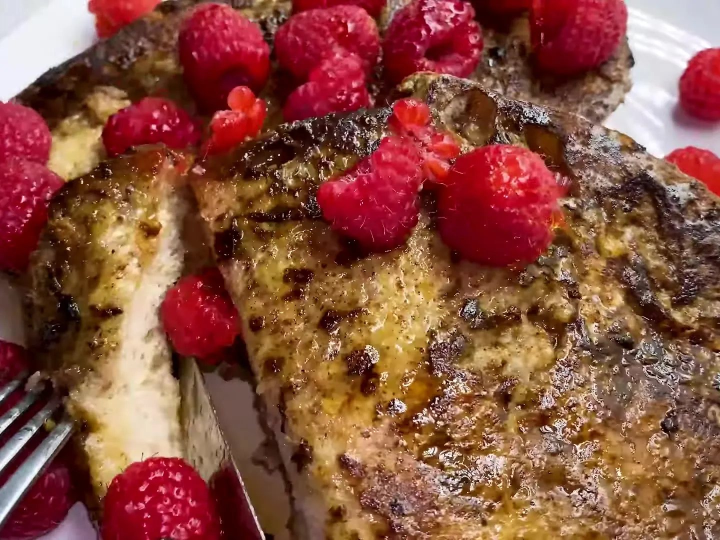 A slice of French toast cut open with a knife and fork to show the soft inside, with raspberries and maple syrup on top