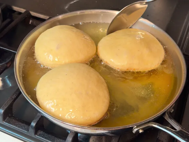 Three pale donuts frying in a stainless steel skillet, with a spoon dipping into the oil to baste them