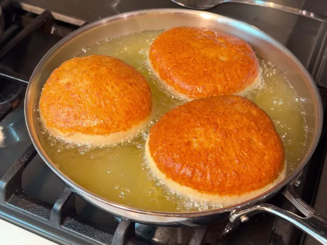 Three deep golden donuts frying in a stainless steel skillet of oil on a gas stove