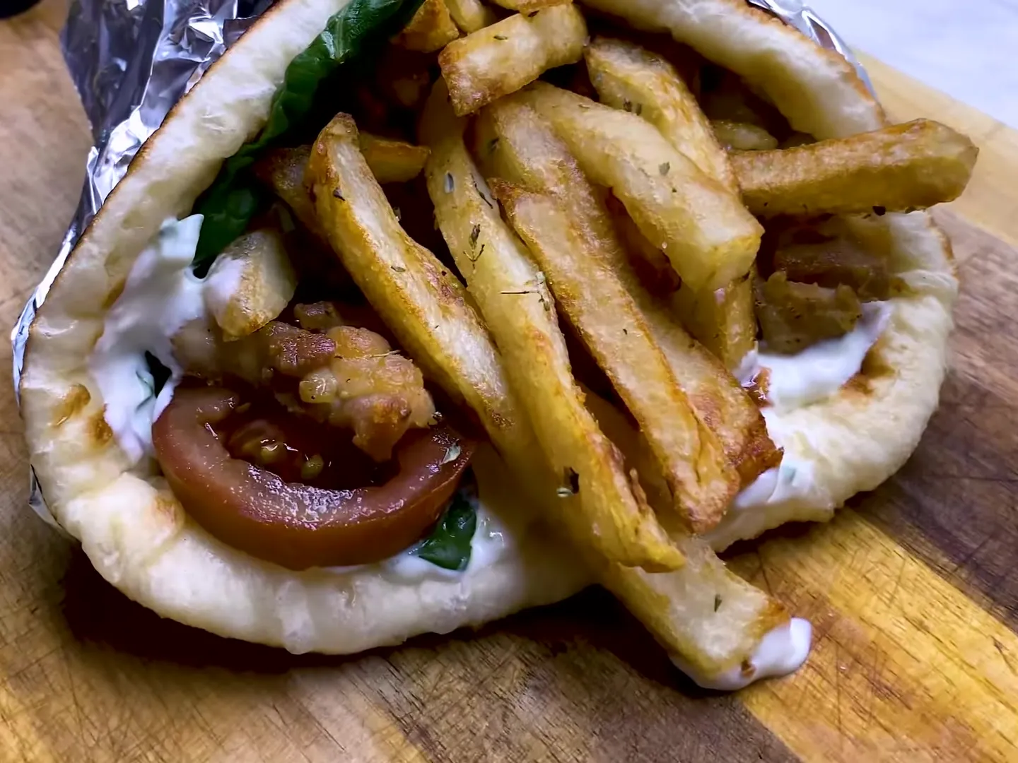 A chicken gyro in homemade pita, half wrapped in foil, with tzatziki, tomato, lettuce and fries, on a wooden board