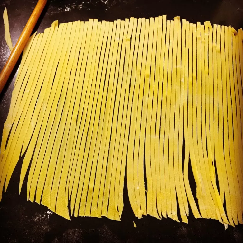 Hand-cut tagliatelle laid out in long strips on a dark, floured counter, next to a wooden rolling pin