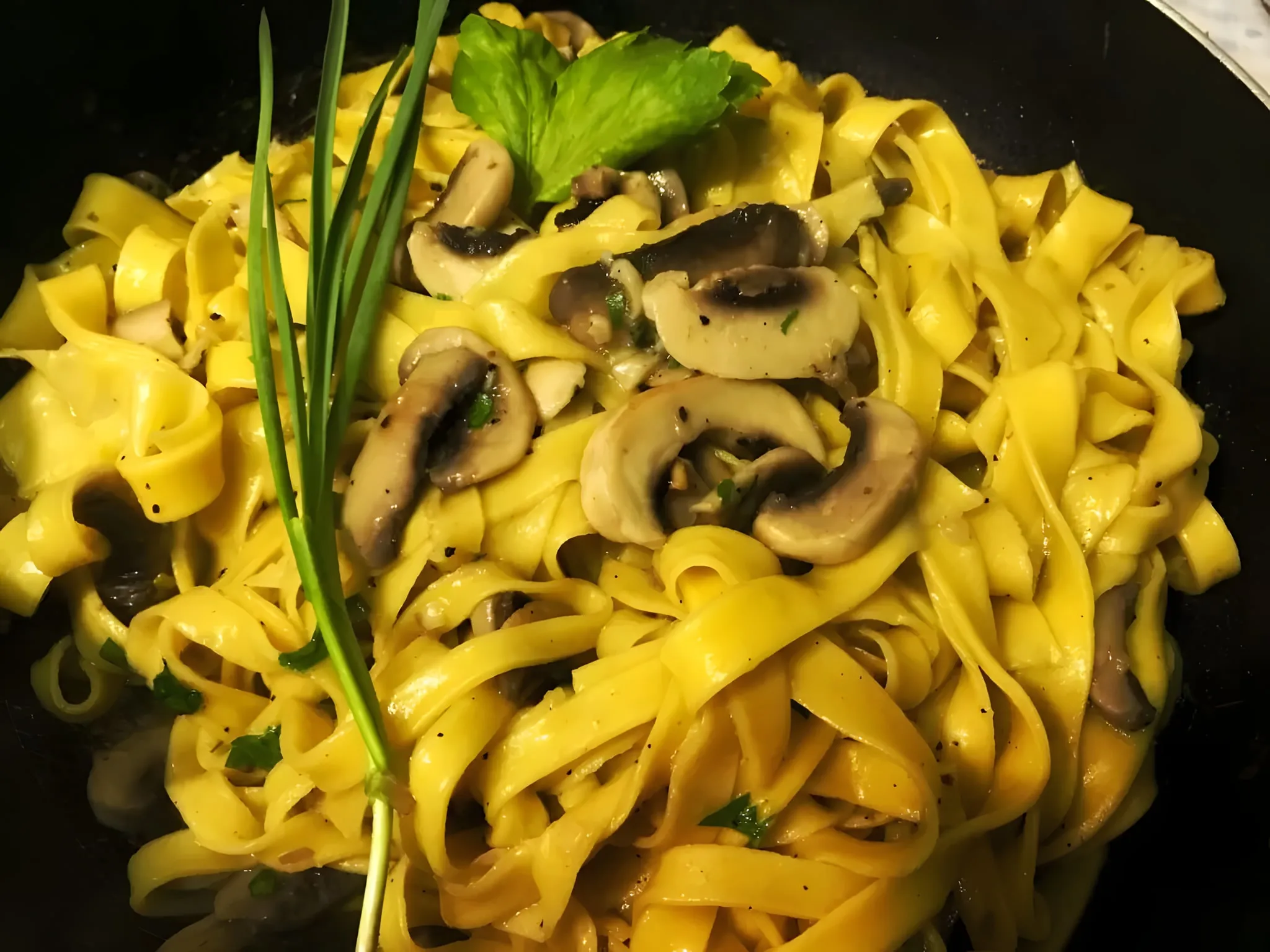 Fresh tagliatelle with sliced white mushrooms and black pepper in a dark pan, with chives and a green herb leaf on top
