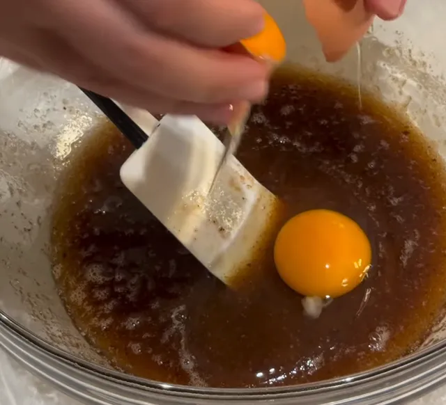 An egg yolk dropping into the brown sugar and butter mixture next to a spatula, with the whole egg already in the bowl
