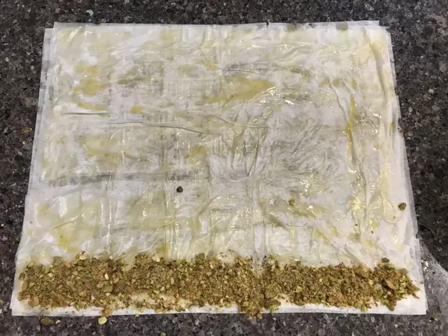 Buttered fillo sheets on a granite counter, with a line of crushed pistachios along the bottom long edge