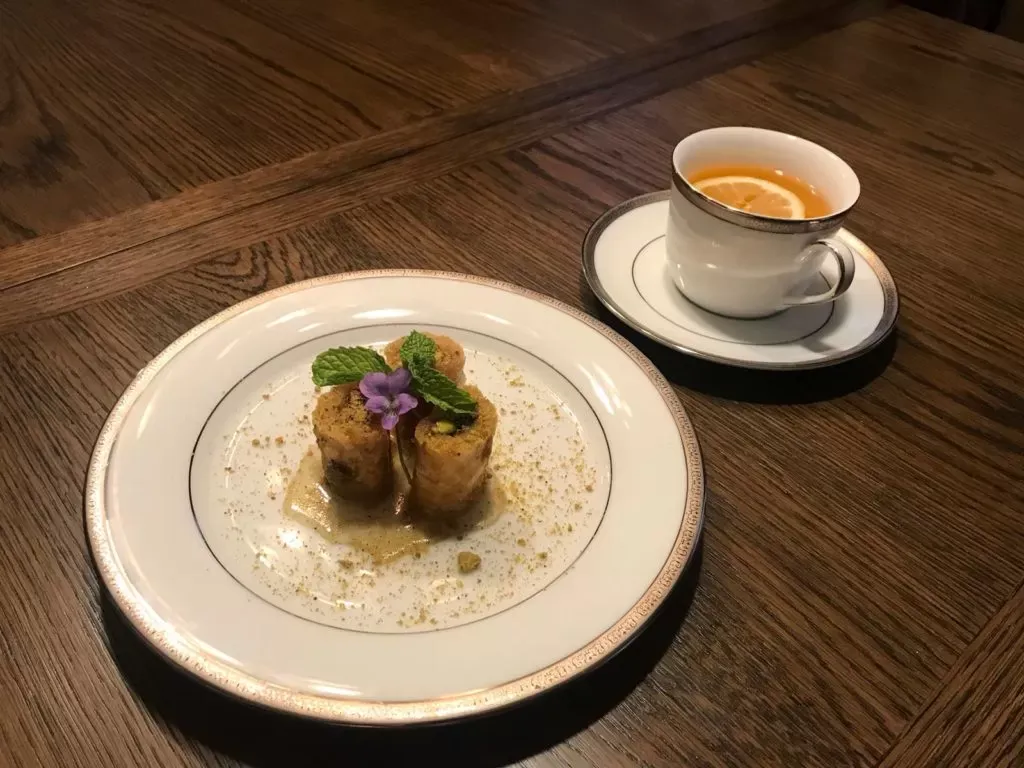 Three pistachio baklava fingers on a white plate with mint leaves and a small purple flower, next to a cup of tea with a slice of lemon, on a wooden table