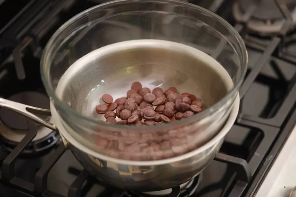 Chocolate chips in a glass bowl set over a saucepan on a gas stove