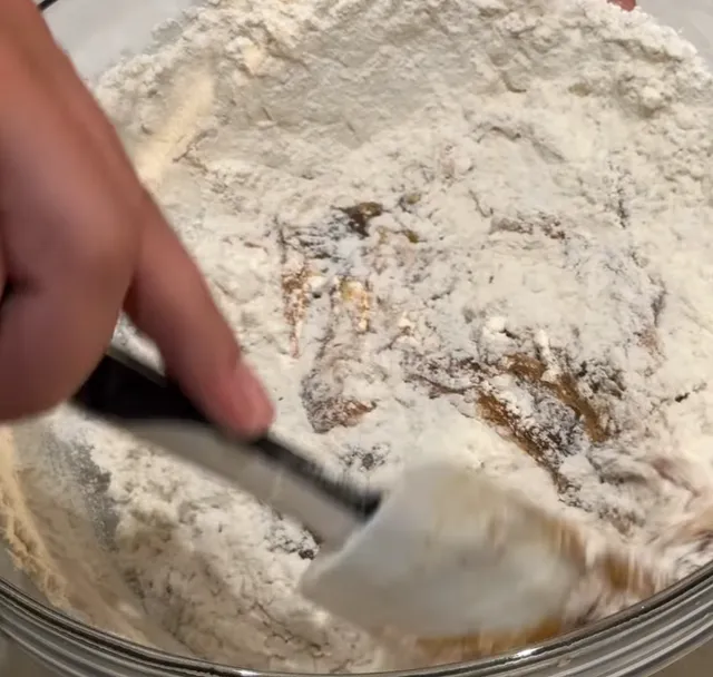 Flour and baking soda being stirred into the brown sugar and butter mixture with a spatula