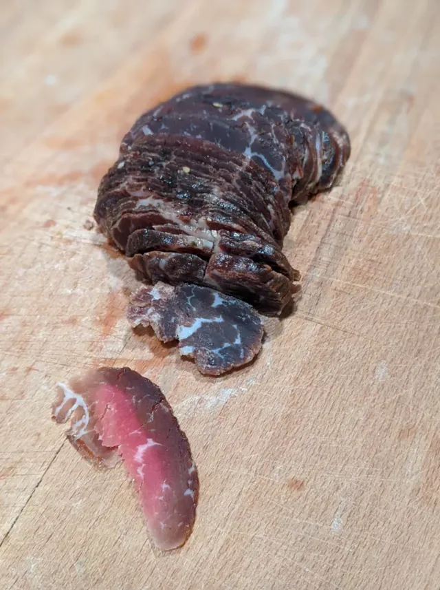 Thin slices of homemade pastirma fanned out on a wooden cutting board, with one slice lying apart to show the deep red inside