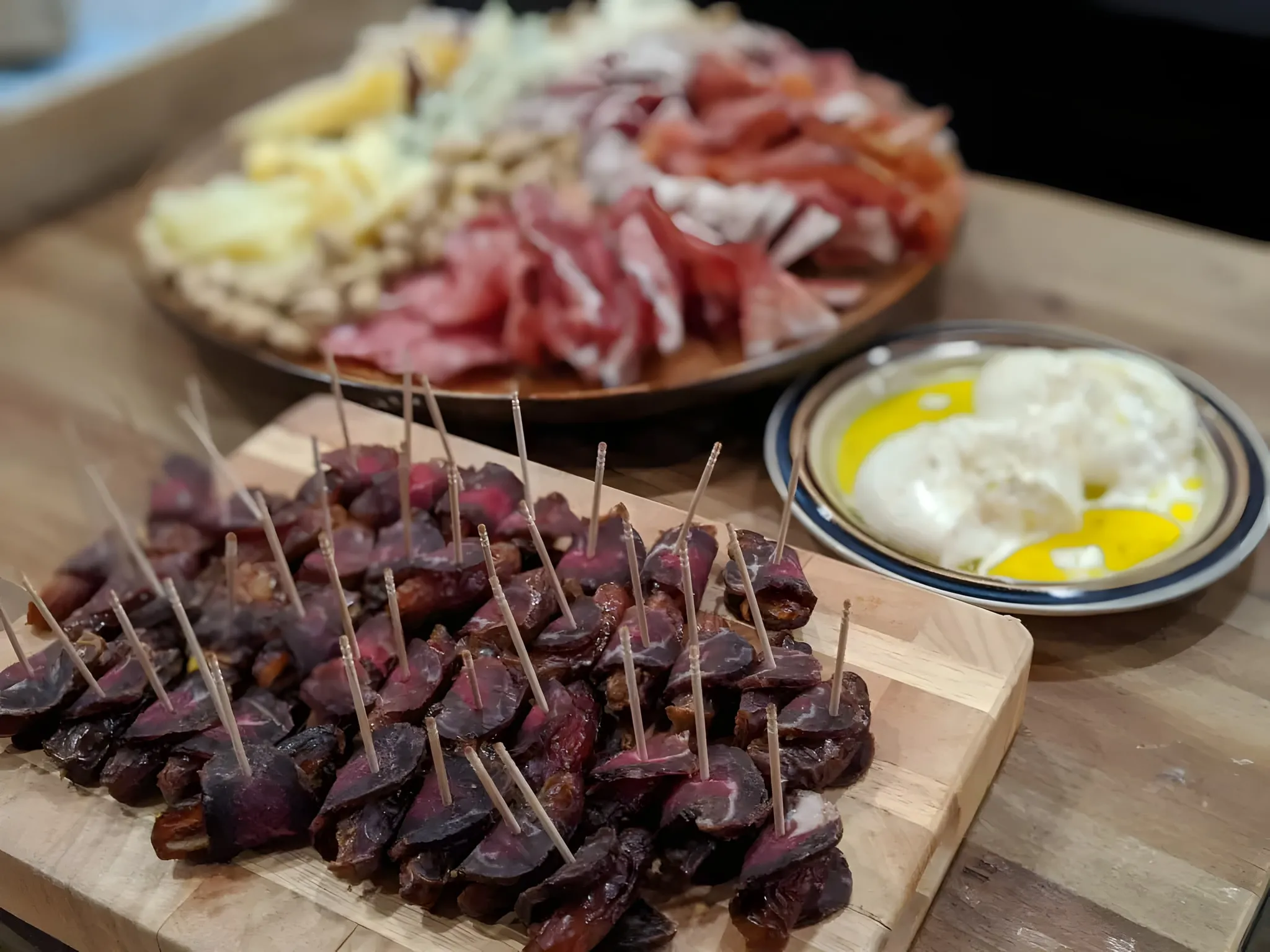 Pitted dates wrapped in thin slices of homemade pastirma, each held with a toothpick, on a wooden board, with a platter of cheese and cured meats and a plate of fresh cheese in olive oil behind