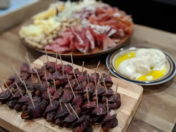 Pitted dates wrapped in thin slices of homemade pastirma, each held with a toothpick, on a wooden board, with a platter of cheese and cured meats and a plate of fresh cheese in olive oil behind