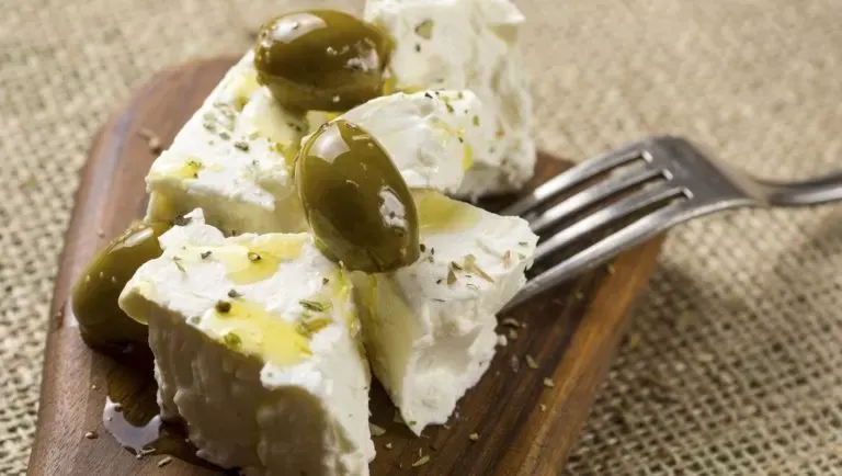 Cubes of white feta with green olives, olive oil and dried herbs on a small wooden board, with a fork