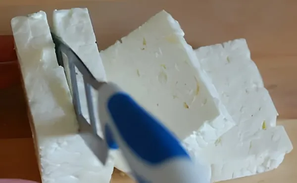 A block of white feta being cut into thick slabs with a blue-handled cheese slicer