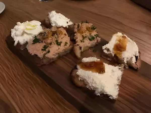 Bruschette on a wooden board, topped with creamy white cheese, some with amber jam and some with a chunky pink spread and parsley