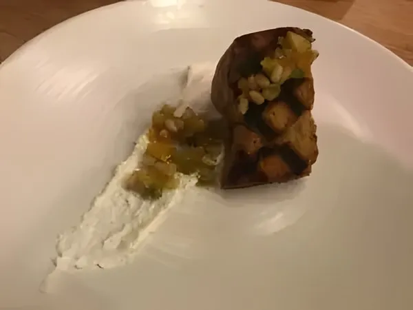 A grill-marked piece of olive cake topped with pine nuts and diced fruit, beside a swipe of white cream on a white plate