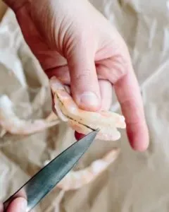 A peeled raw shrimp held between the fingers while a small knife cuts along its back