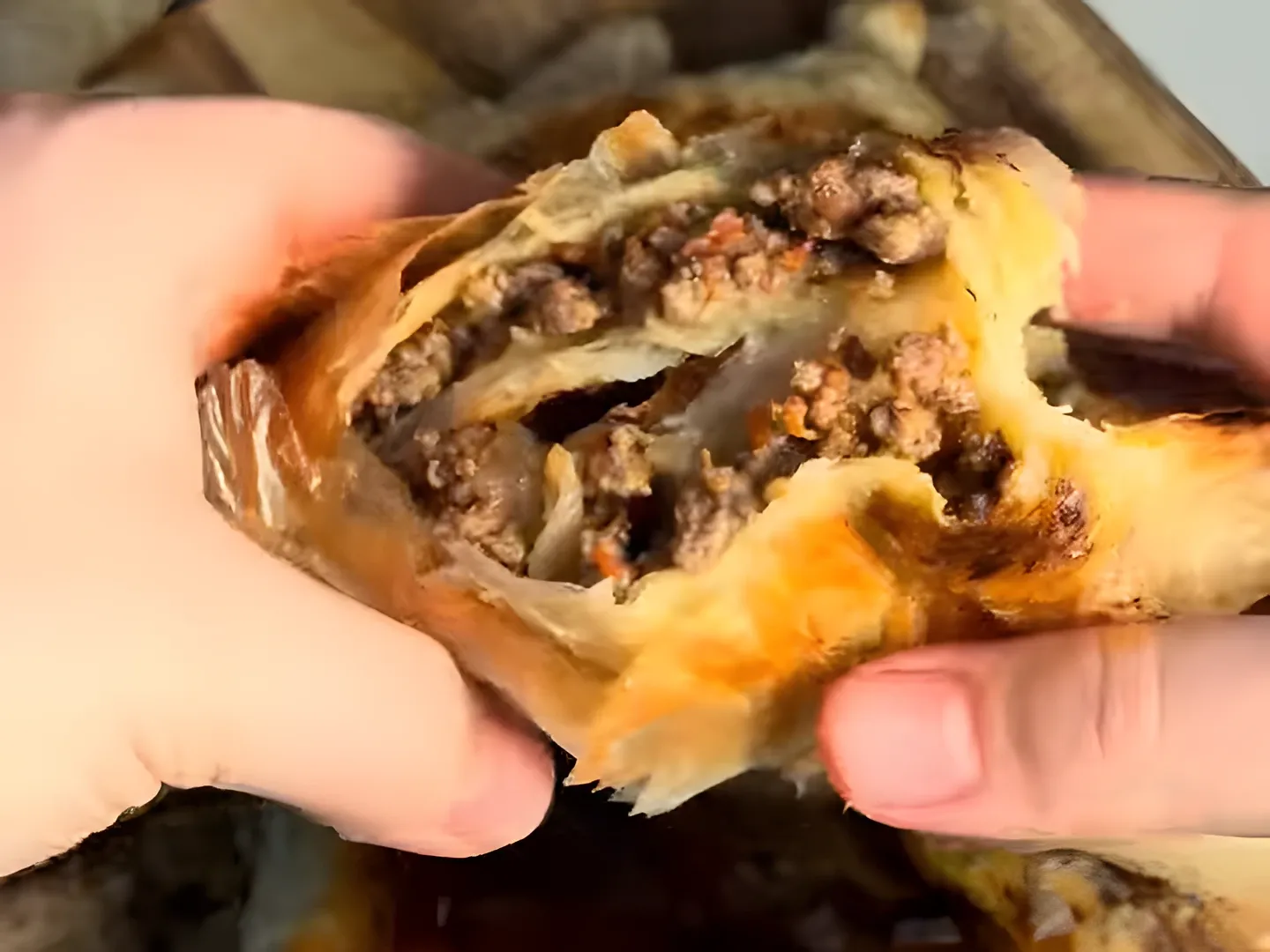 Two hands pulling a baked byrek triangle apart, showing the thin layers and the beef filling inside