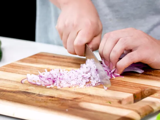 Hands chopping thin slices of red onion into small pieces on a wooden board, next to a pile of finely chopped onion