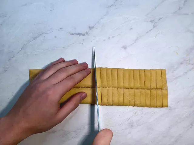 A hand holding a folded sheet of pasta dough while a knife cuts it into even strips on a marble counter