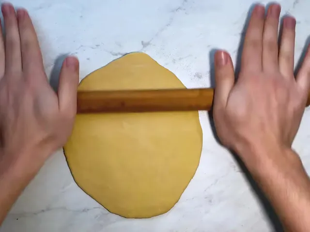 Two hands rolling a round yellow sheet of pasta dough with a long, thin wooden rolling pin on a marble counter