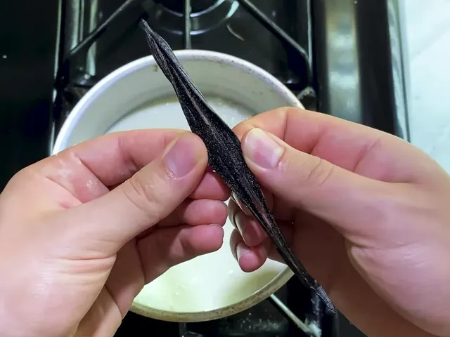 Hands holding a split vanilla bean over a saucepan of milk on the stove