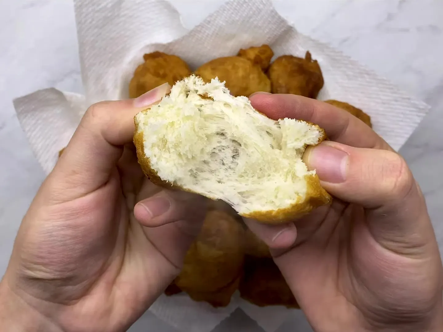 Hands pulling a petulla apart to show the soft, stretchy inside, over a bowl of golden petulla