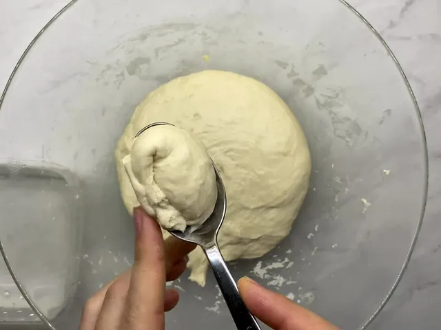 A spoonful of sticky petulla dough lifted from the bowl, with a dish of water beside it for the spoon