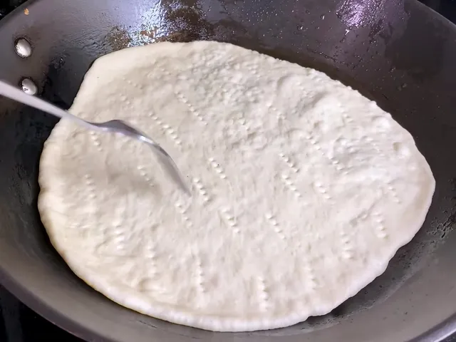 A fork poking rows of holes in a raw pita as it cooks in a frying pan
