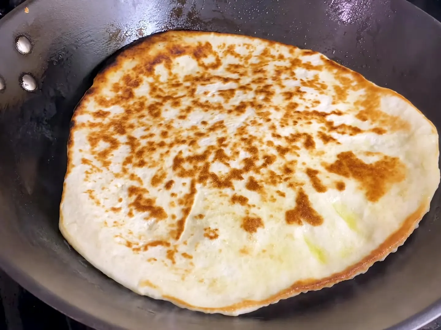A homemade Greek pita in a dark frying pan, golden brown on top after flipping