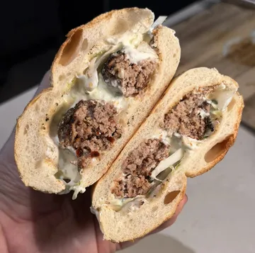 Bukë me qofte, a bread roll with grilled qofte, tzatziki and onions, cut in half and held in the hand