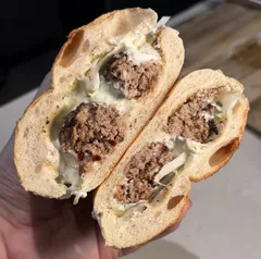 Bukë me qofte, a bread roll with grilled qofte, tzatziki and onions, cut in half and held in the hand