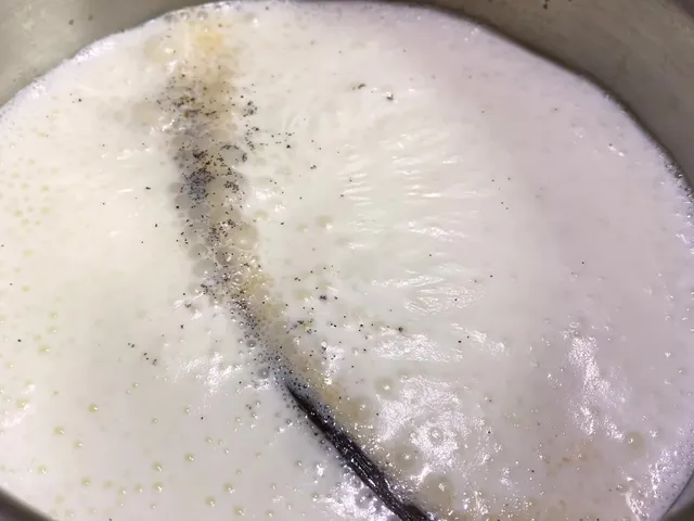 Milk foaming in a saucepan around a split vanilla bean, with the seeds spreading through the milk