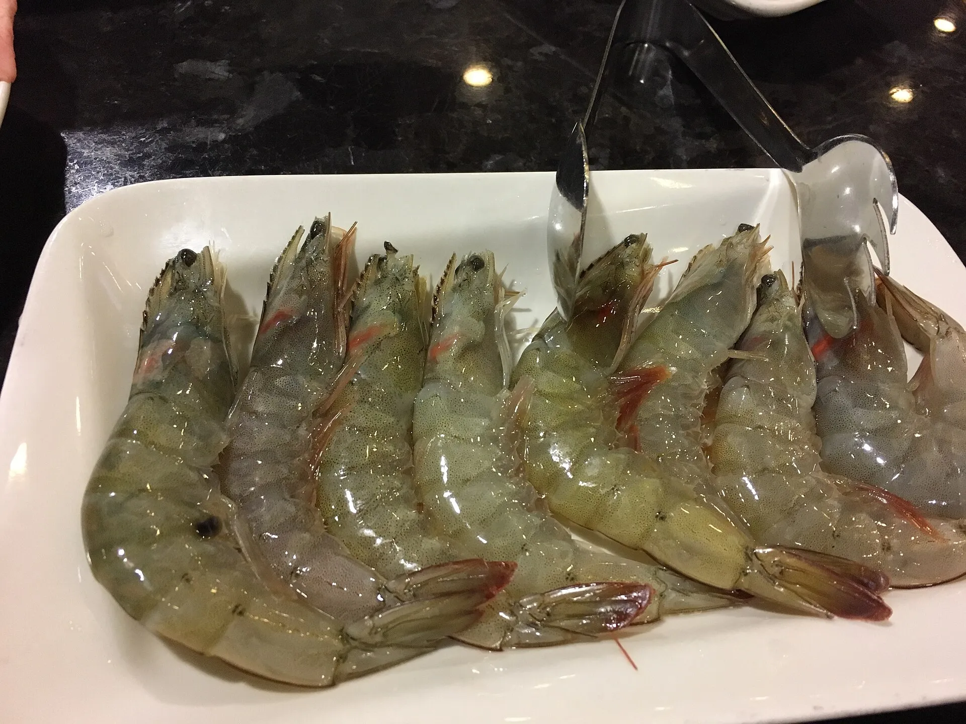 Raw whole shrimp with their heads and shells on, lined up in a white dish, with serving tongs