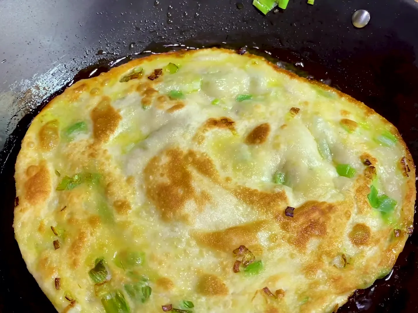 A scallion pancake frying in a dark pan, golden brown in patches, with bits of green scallion