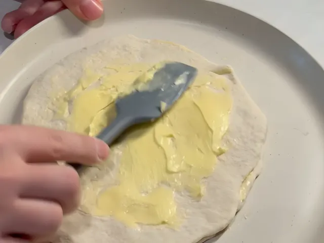 Spreading soft butter over a round sheet of dough on a plate with a grey silicone spatula
