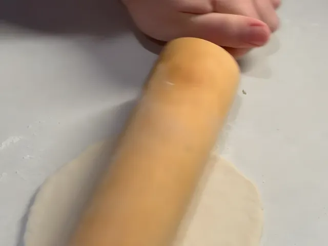 A wooden rolling pin rolling a ball of dough into a thin round sheet