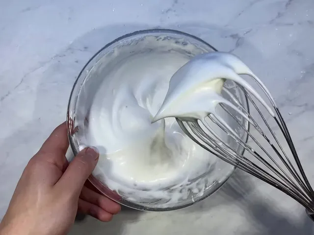 Stiff, glossy egg whites holding a peak on a whisk over a glass bowl