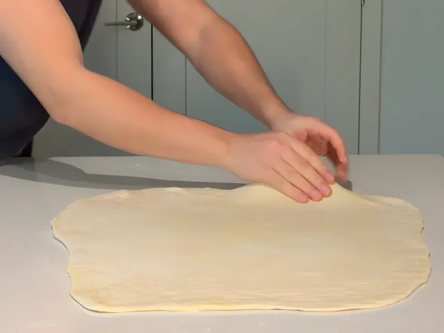 Pressing out the edge of the rolled stack of byrek dough, now a large rectangle, on a kitchen island