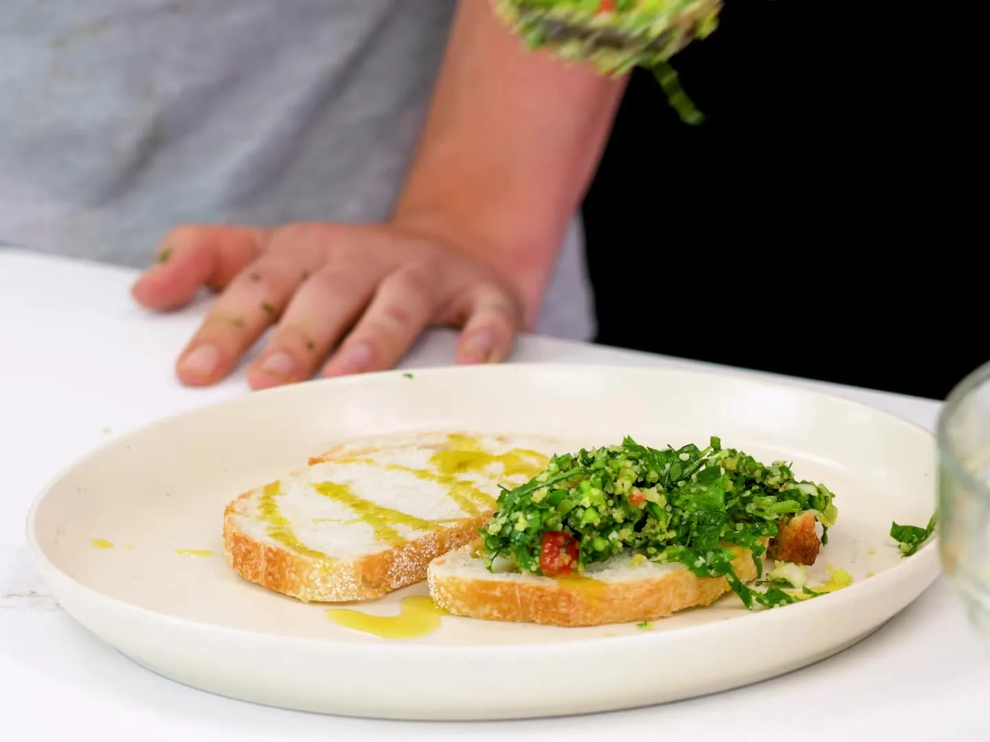 Two slices of bread drizzled with olive oil on a white plate, one topped with tabbouleh, finely chopped parsley with bulgur and bits of tomato