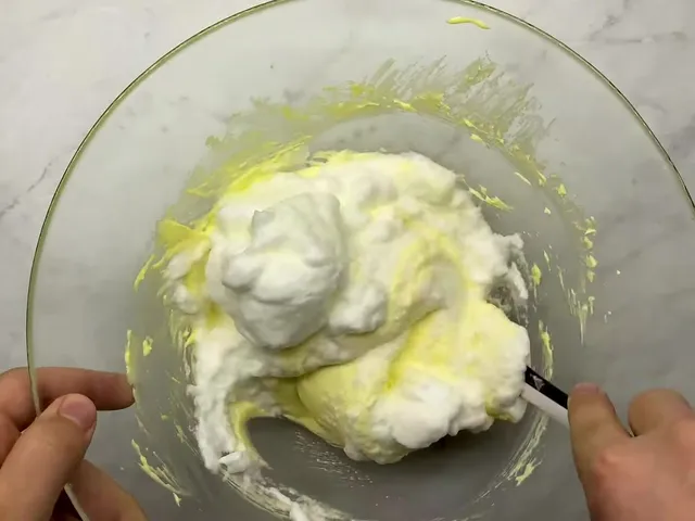 Whipped egg whites being folded into the yellow mascarpone cream with a spatula in a glass bowl