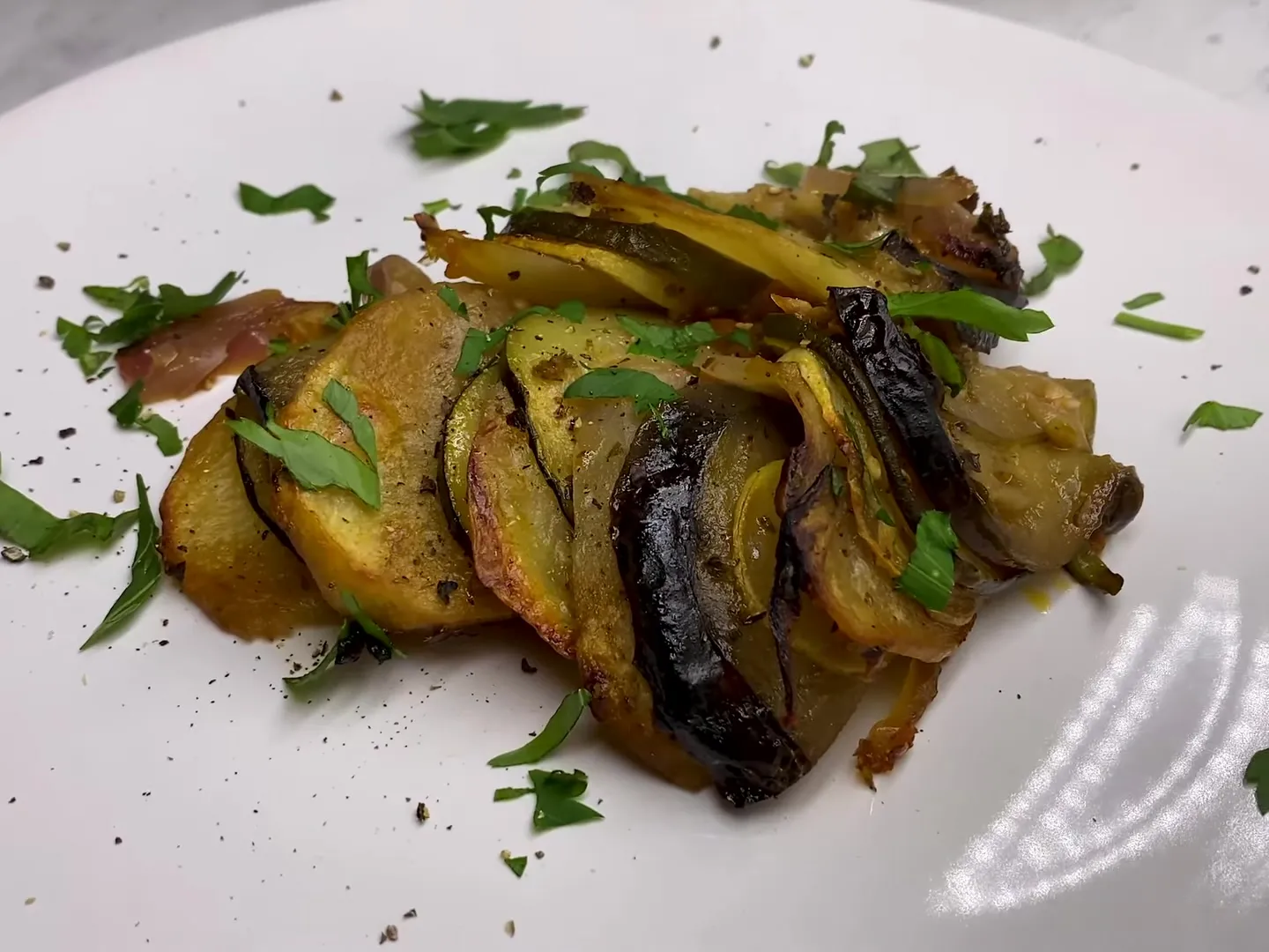 A serving of turli on a white plate, overlapping slices of potato, eggplant and squash with chopped parsley