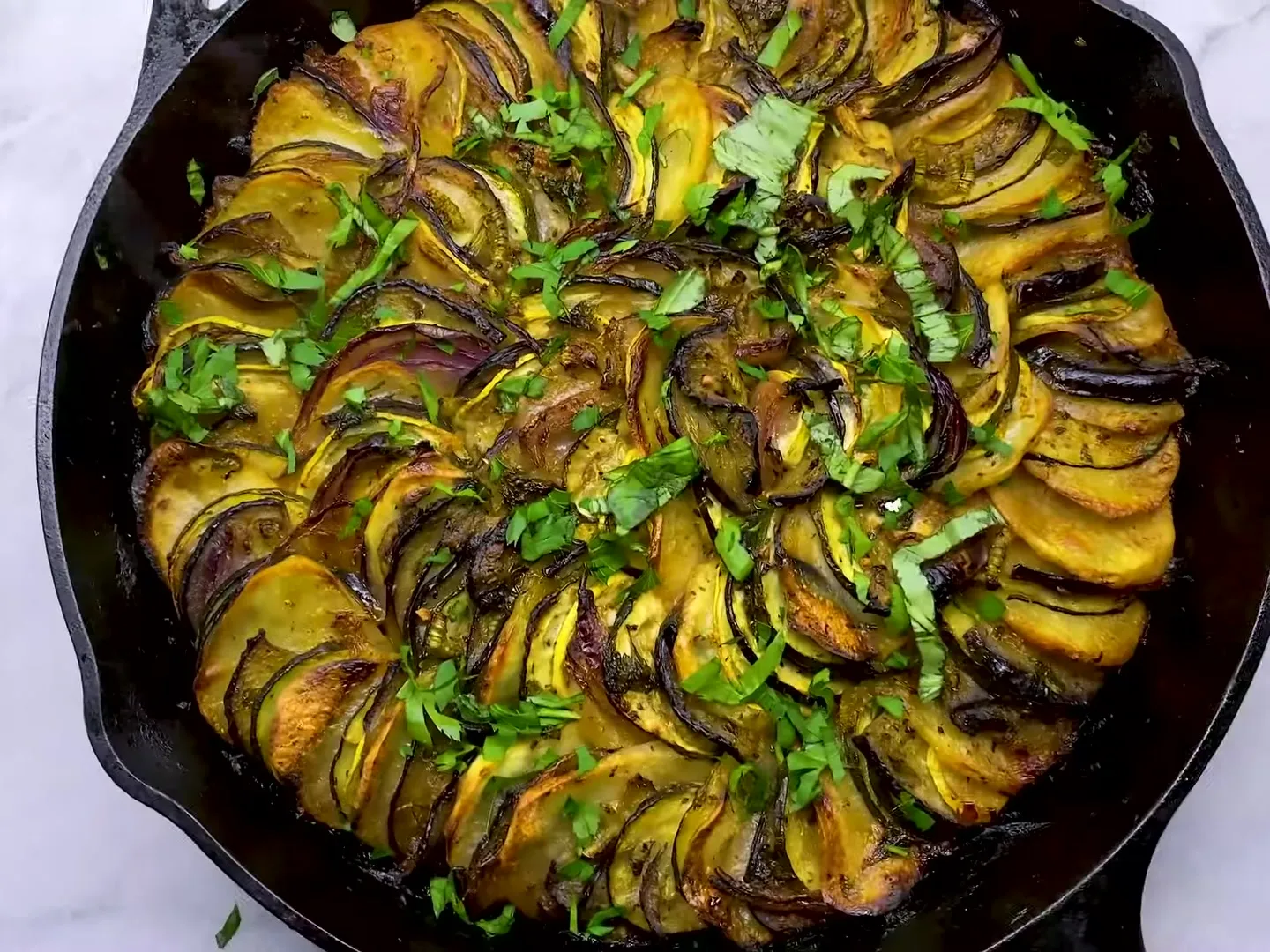Turli in a black cast-iron skillet, thin slices of potato, eggplant, zucchini, yellow squash and red onion baked golden in a spiral, with parsley and basil on top