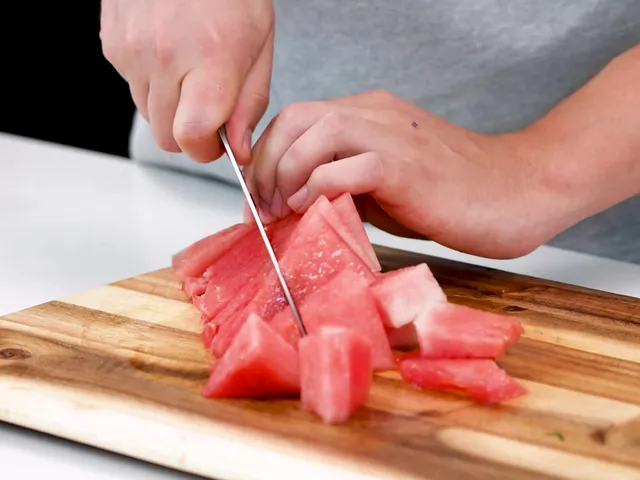 Hands cutting a strip of watermelon into cubes on a wooden board