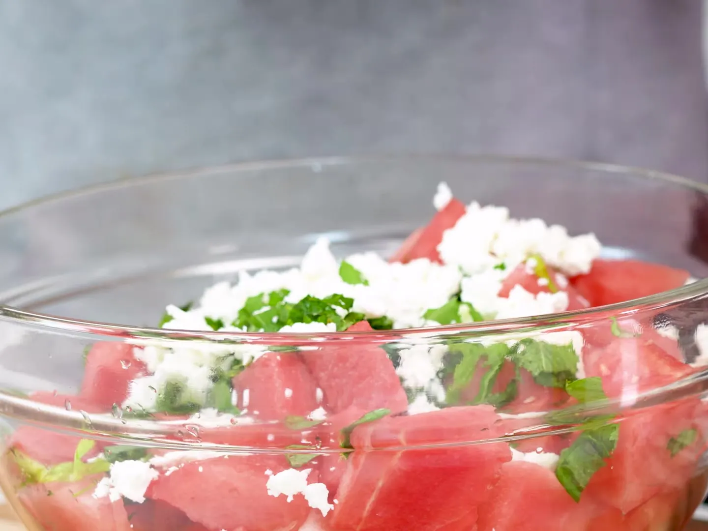 A glass bowl of watermelon cubes topped with crumbled feta and strips of fresh basil and mint